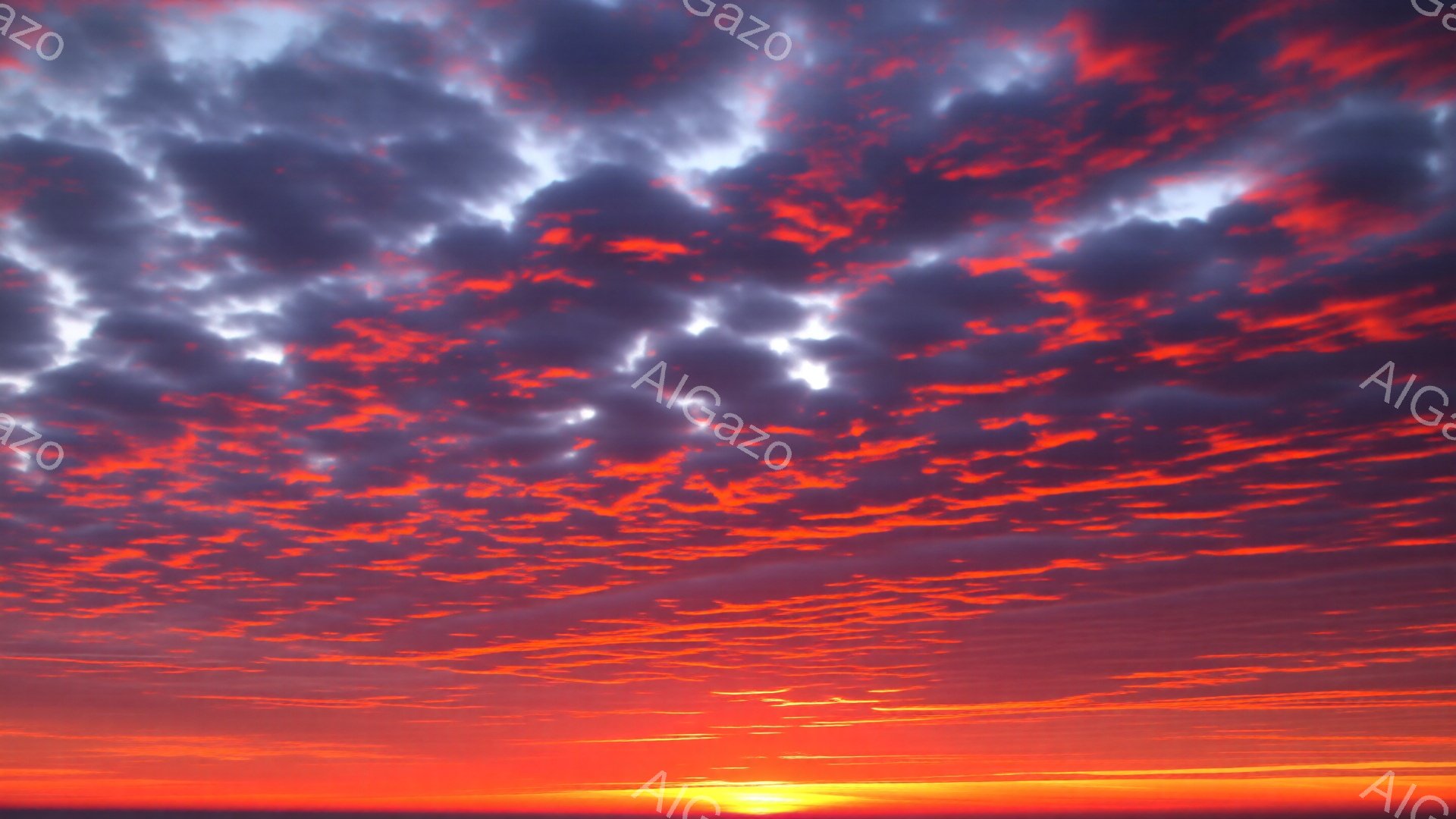 空は燃えるような赤とオレンジ色で染まり、まるで炎が広がっているようだ。雲は独特の縞模様を描き、その下には地平線に沿って薄い暗い帯が見える。全体として、力強くドラマチックな夕焼けの雰囲気で、息をのむような美しさだ。