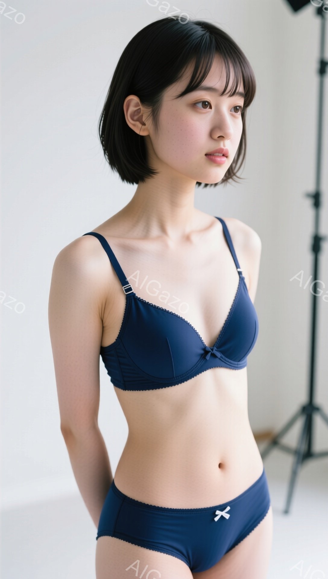 暗い青色のブラとショーツを着用した若い女性が写っています。彼女は肩までの長さの黒髪で、顔は静かで穏やかな表情をしています。背景は白く、スタジオの機材の一部が見え、全体的に清潔でミニマルな雰囲気が漂って - AI生成フリー素材