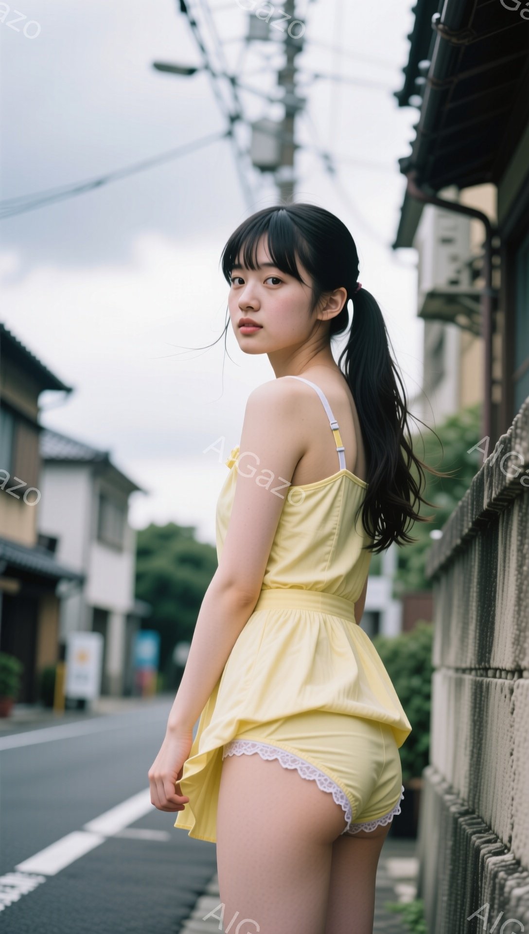 黒髪をポニーテールにした若い女性が、薄黄色のワンピースを着て、路肩に立っています。彼女は少し振り返り、どこか寂しげな表情をしています。背景には古い木造建築が並び、曇り空の下、どこか懐かしい雰囲気の街並みが広がっています。