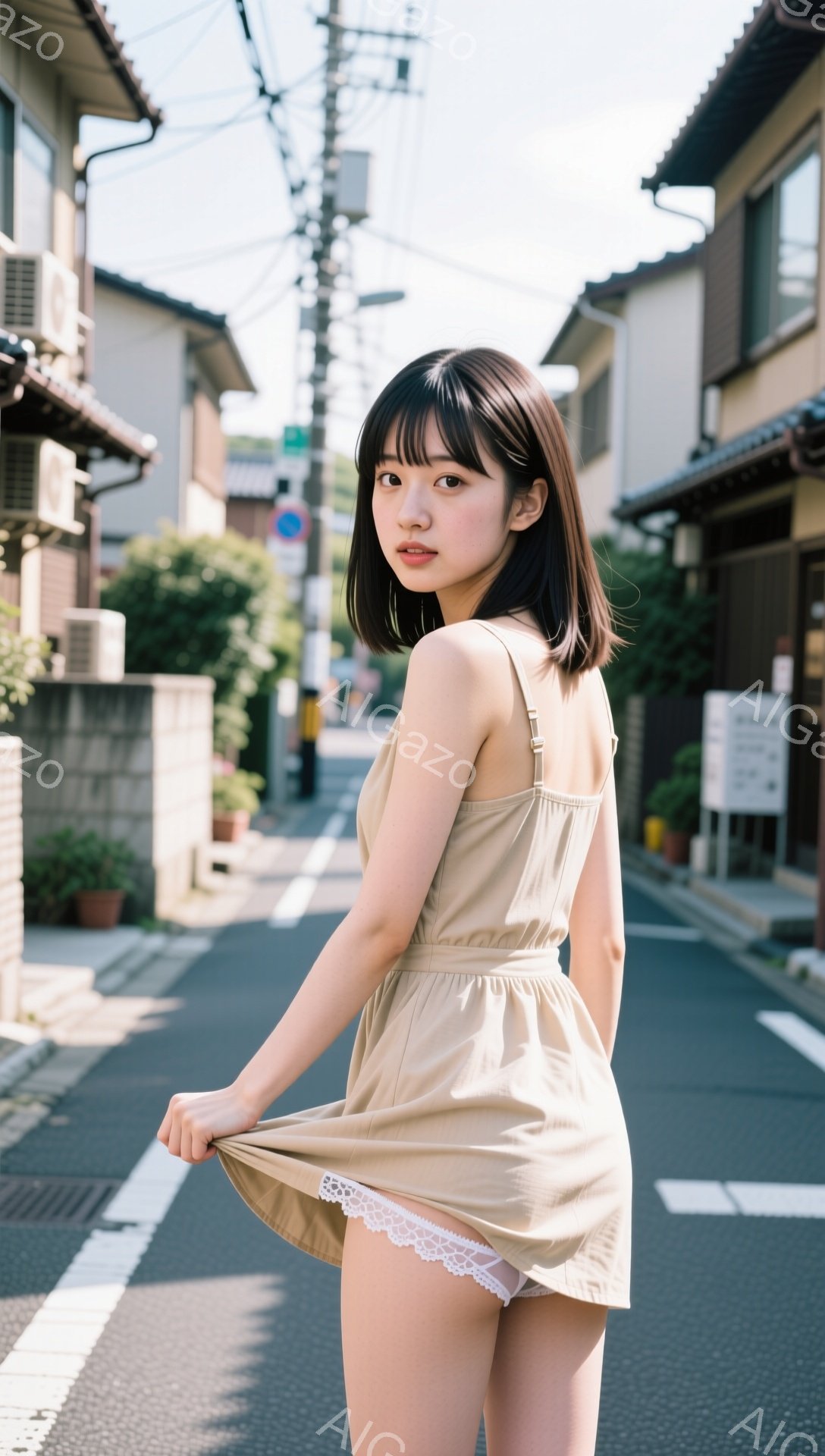 浅いベージュ色のキャミソールワンピースを着た若い女性が、スカートの裾を少し持ち上げて立っている。彼女は黒髪のショートボブで、少し微笑んでこちらを見ている。背景は日本の住宅街の狭い道で、両脇に木造の建物が並び、日差しが強く、夏の穏やかな雰囲気である。