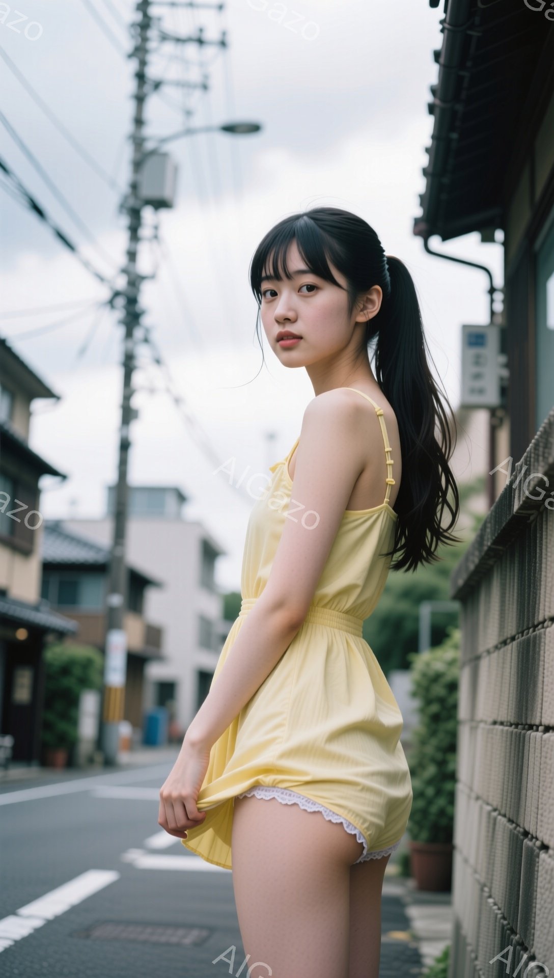 若い女性が黄色いミニワンピースを着て、路地裏に立っている。彼女は黒いロングヘアをポニーテールにし、少し微笑んでこちらを見ている。背景には、コンクリートの壁、植物、住宅が見え、曇天の空が広がり、静かで落ち着いた雰囲気が漂う。