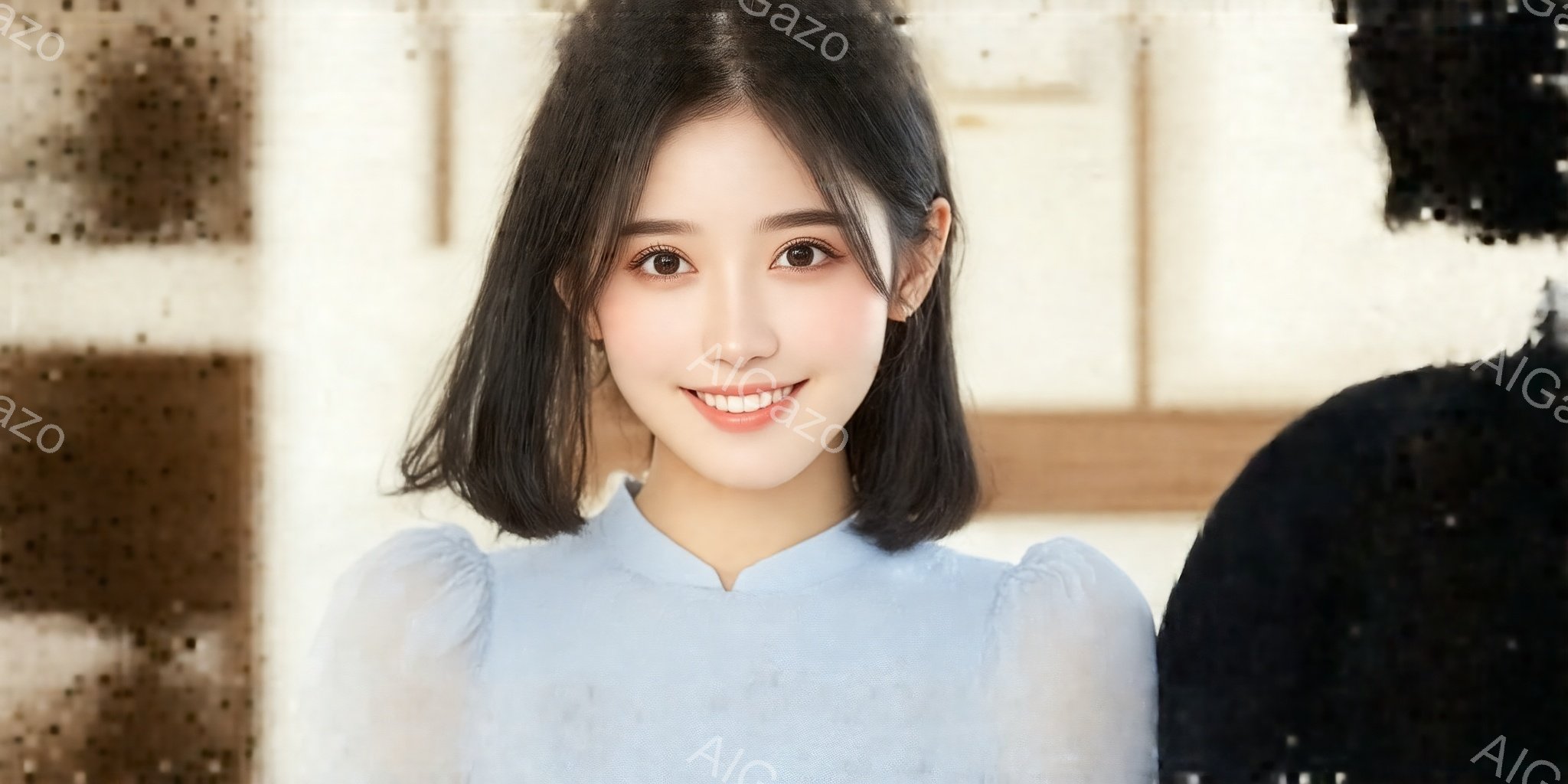 ショートヘアの女性が微笑んでいます。彼女は淡い水色のブラウスを着ており、顔色は明るく、表情は穏やかです。背景はぼやけており、黒いものがわずかに見えますが、女性に焦点が当てられています。