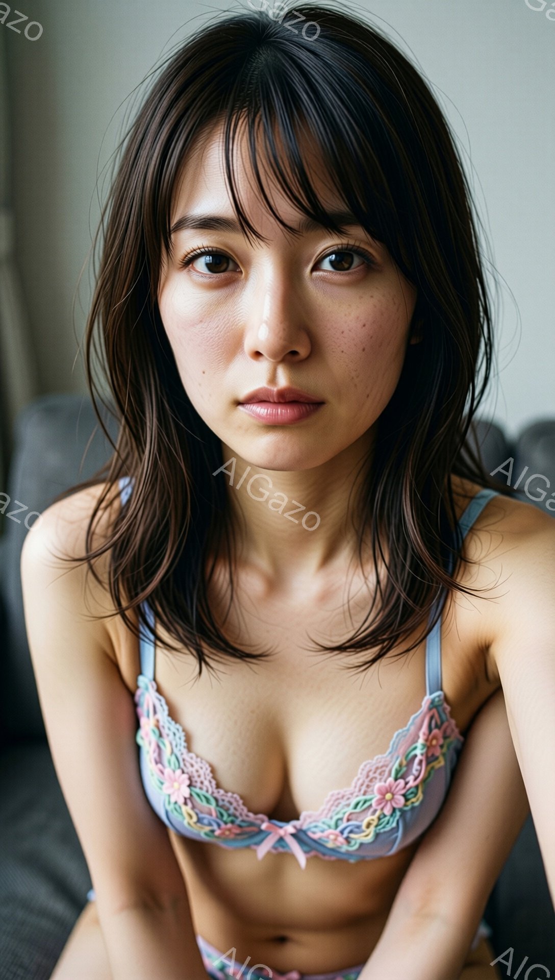 長い黒髪の女性が、淡いピンクと水色のレースのブラレットとショーツを身につけて座っています。彼女はカメラの方を少し物憂げな表情で見ており、その顔にはそばかすが点在しています。背景はぼやけたグレーのソファで、全体的に柔らかく、親密な雰囲気を醸し出しています。