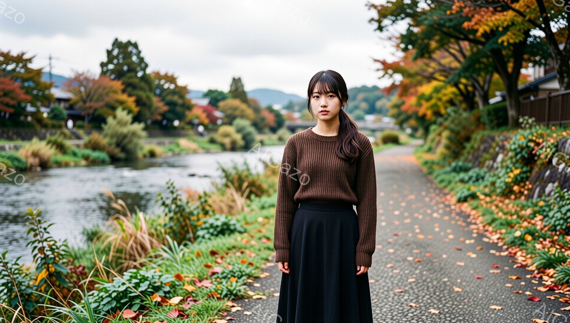 長い黒髪をセンターパートにした若い女性が、茶色のセーターと黒のロングスカートを着て立っている。彼女は表情は穏やかで、少し物憂げな様子を見せている。背景には、紅葉が美しい川沿いの道が広がり、秋の落ち着いた雰囲気が漂っている。